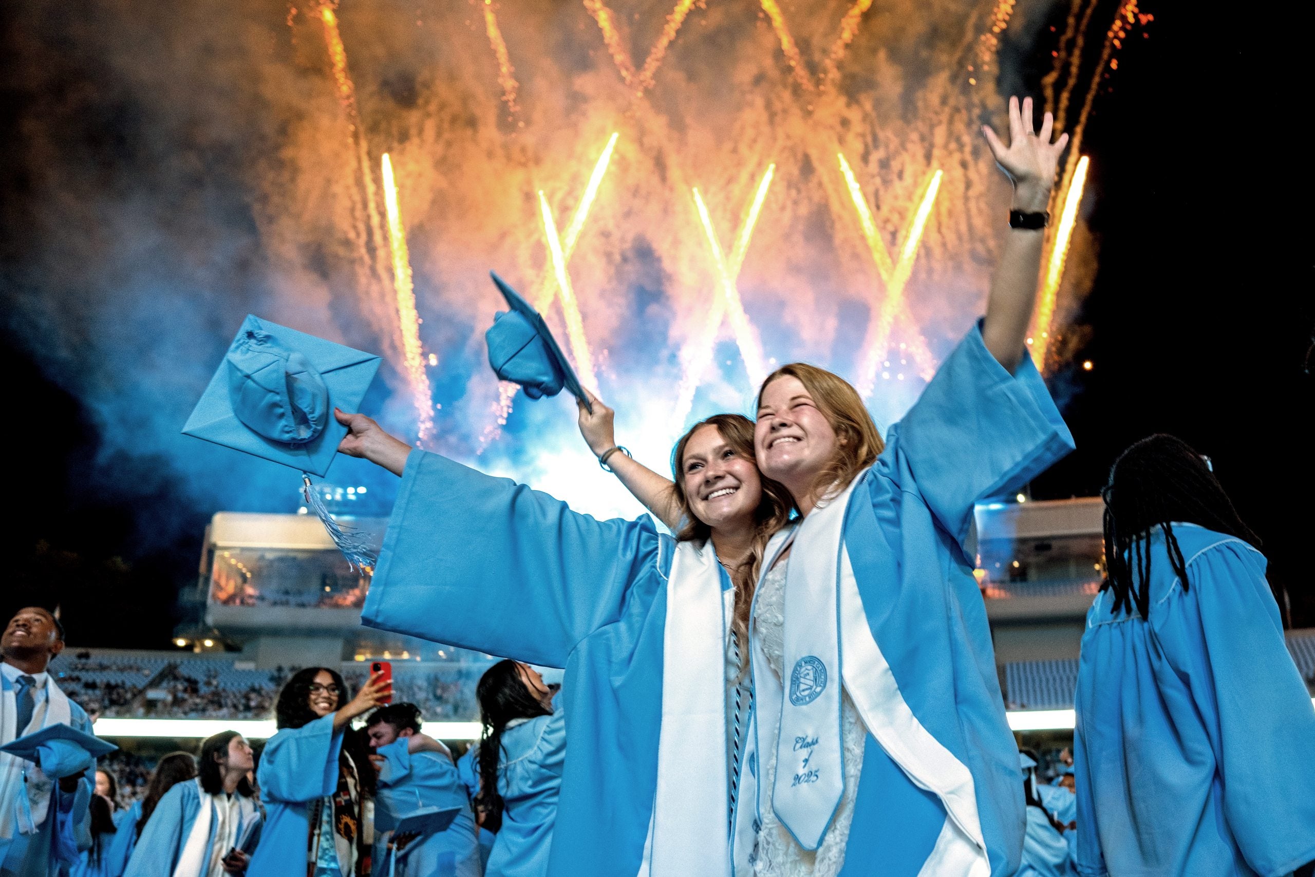 Graduates hug and hold their caps in the air as fireworks go off behind them at Spring Commencement.