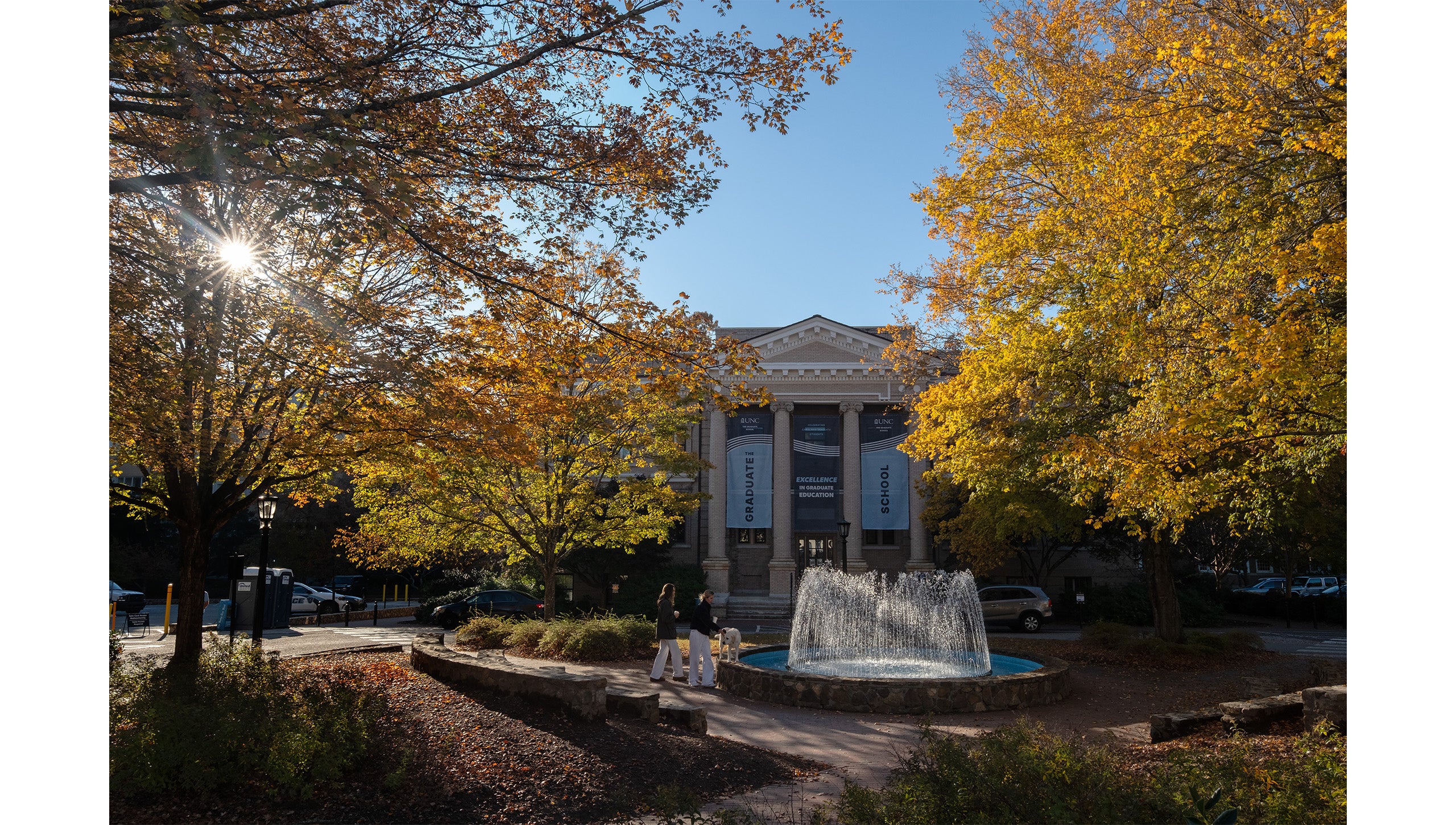 Two people and a dog gathered by Fordham Fountain outside Bynum Hall on the campus of UNC-Chapel Hill on a sunny fall day with trees with yellow, orange and green leaves also pictured.