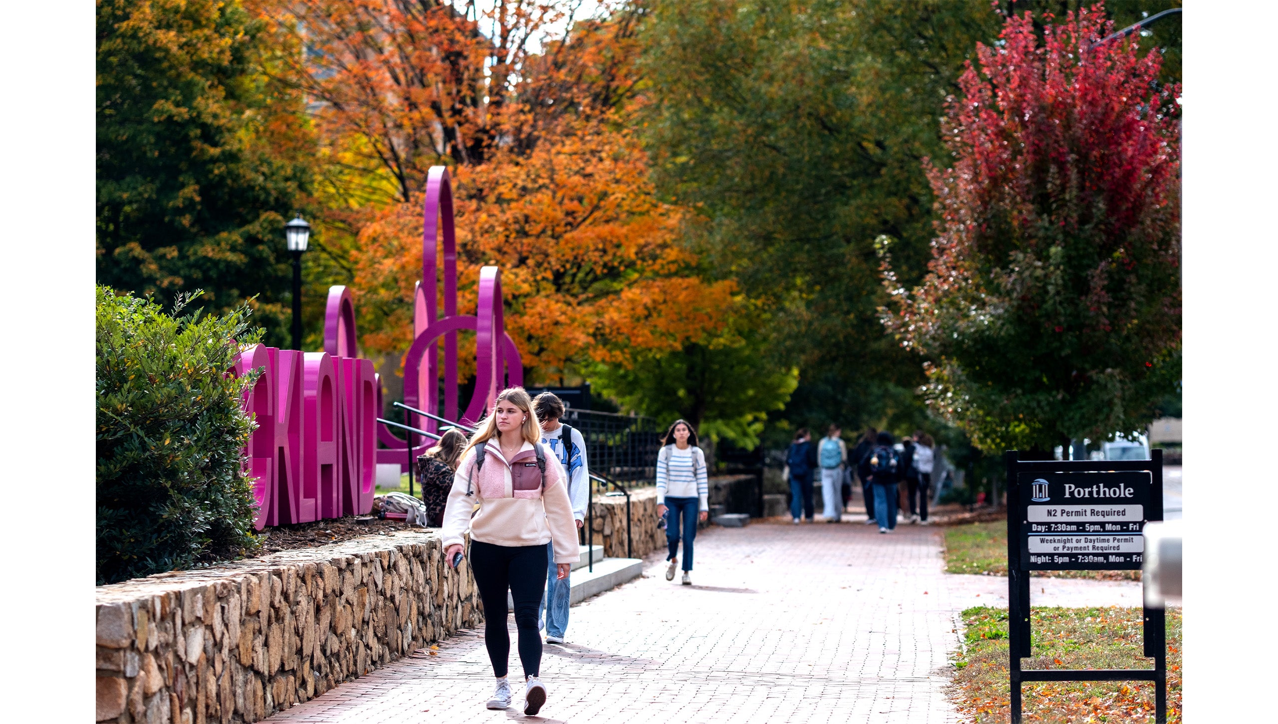 UNC-Chapel Hill students walking down a brick pathway near the Ackland Art Museum. In the background are trees with green, yellow, orange and red leaves.