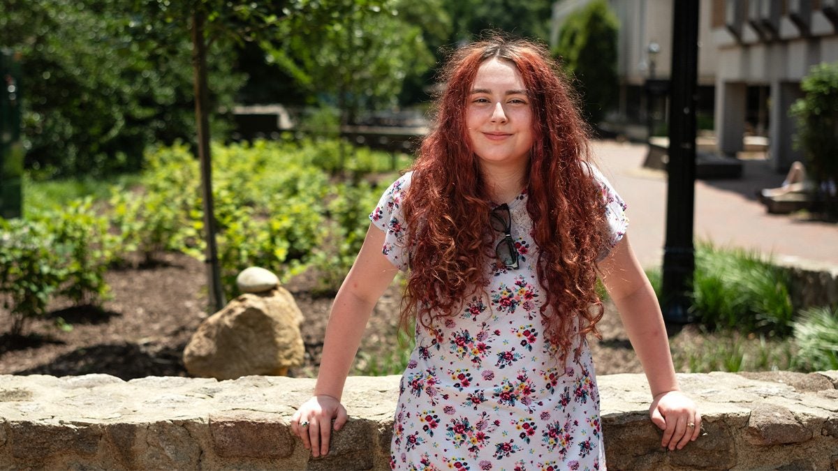 Zoey Ainsworth posing for a portrait standing up and leaning back against a stone ledge on the campus of UNC-Chapel Hill on a sunny afternoon.