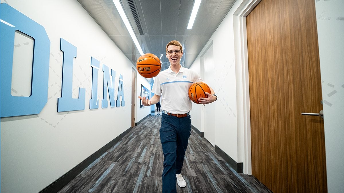 James Blizzard holding two basketballs walks down a hallway smiling at the camera.