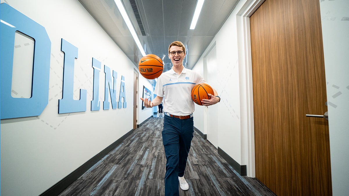 James Blizzard holding two basketballs walks down a hallway smiling at the camera.