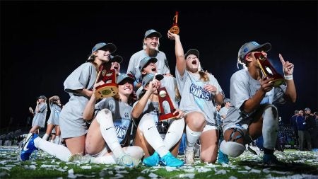 UNC women's soccer players celebrating as a group after national title on field. Players are wearing national championship T-shirts over their jerseys and hats, and one player is holding the trophy.