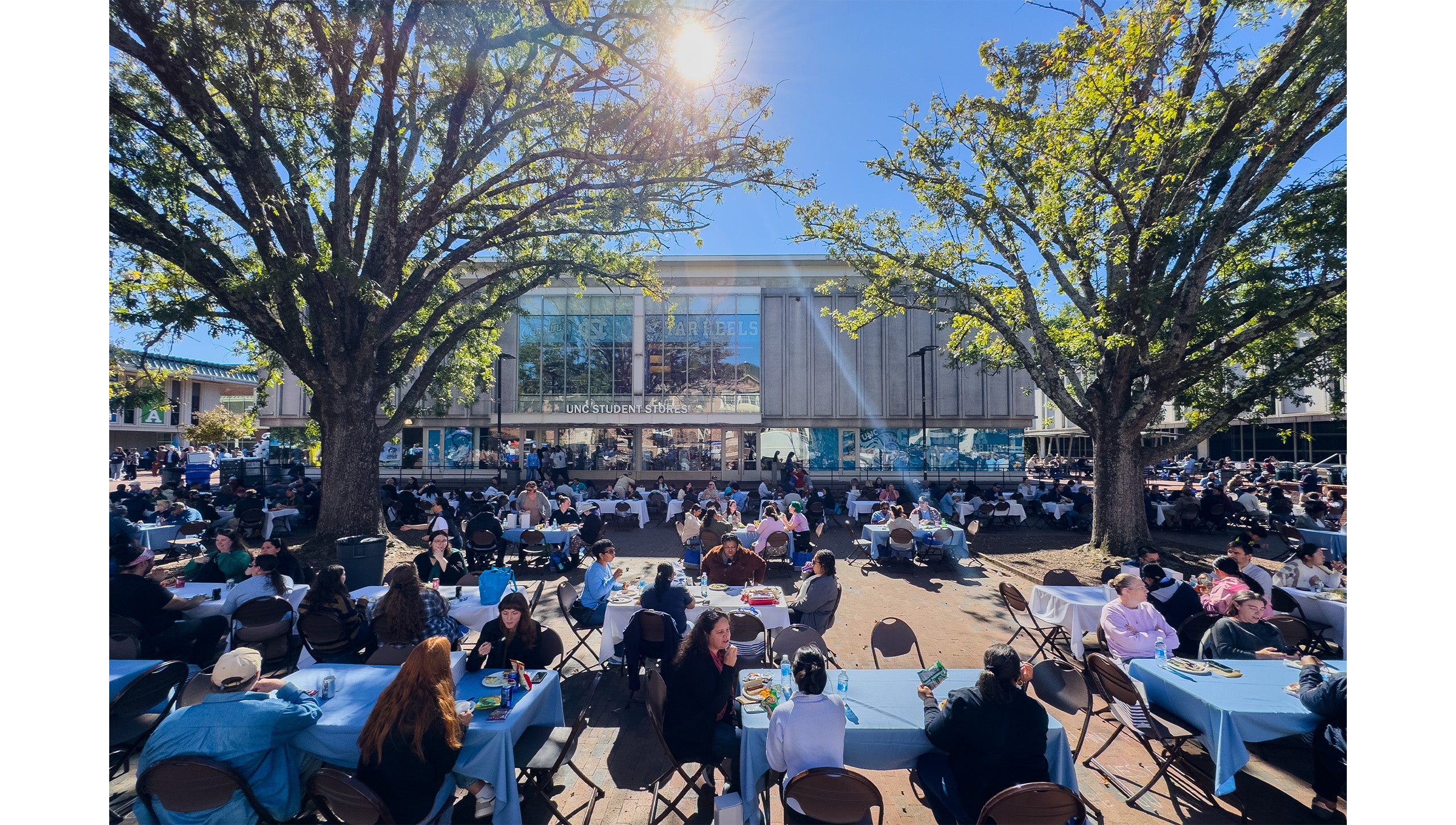 Wide-angle shot of many UNC-Chapel Hill employees eating lunch at tables set up in the Pit in front of Student Stores on a sunny day.