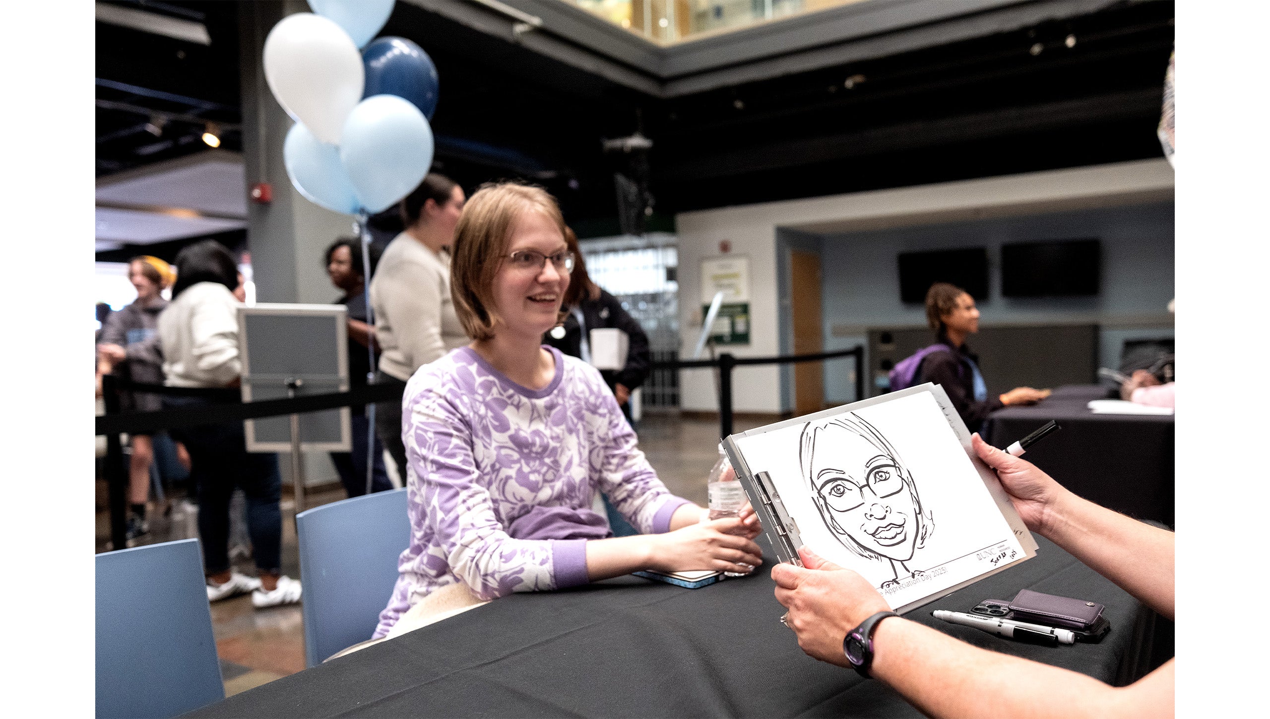 A woman sitting at a table and having her picture drawn by an artist.