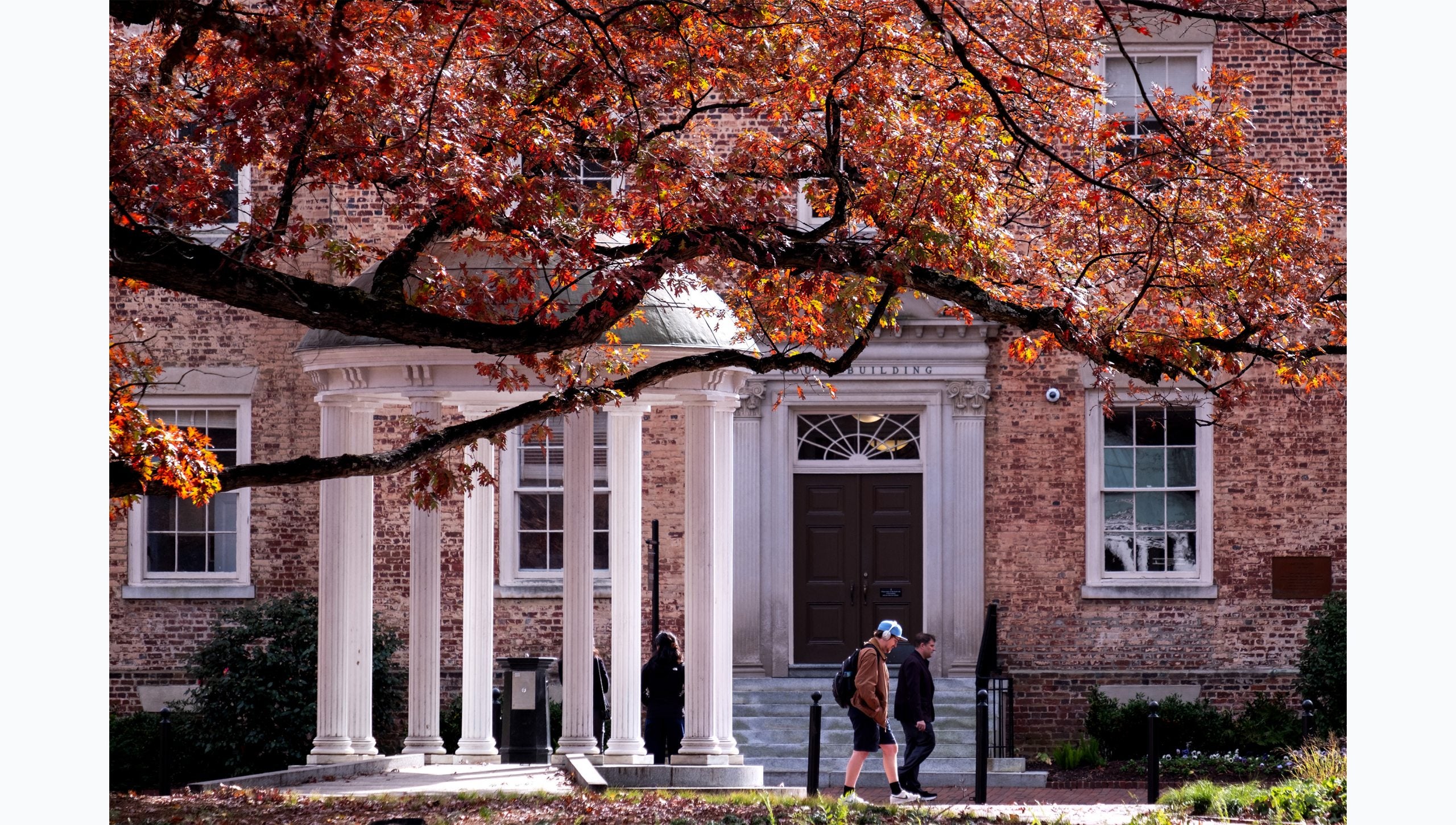People walking by the Old Well on the campus of UNC-Chapel Hill on a fall day. Branches and yellow, red and orange leaves are seen hanging in the foreground.