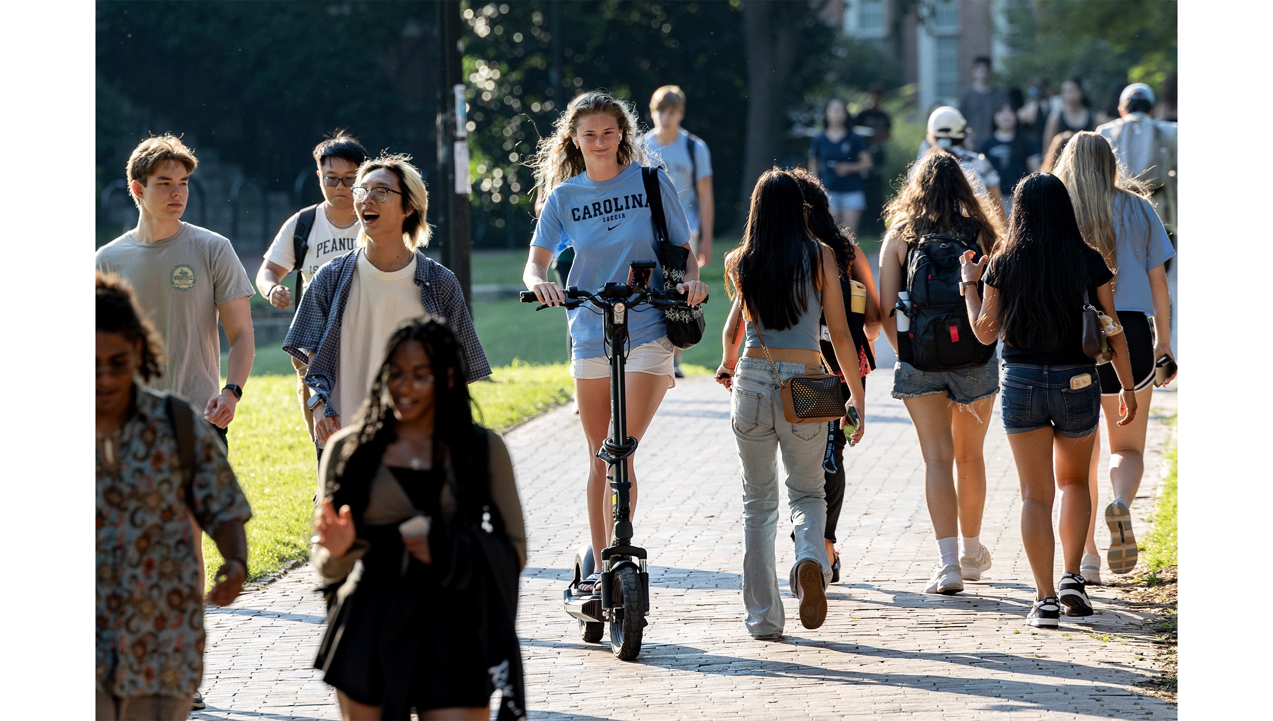 Crowd of UNC-Chapel Hill students walking on brick pathway on Polk Place as a student rides an electric scooter between students.