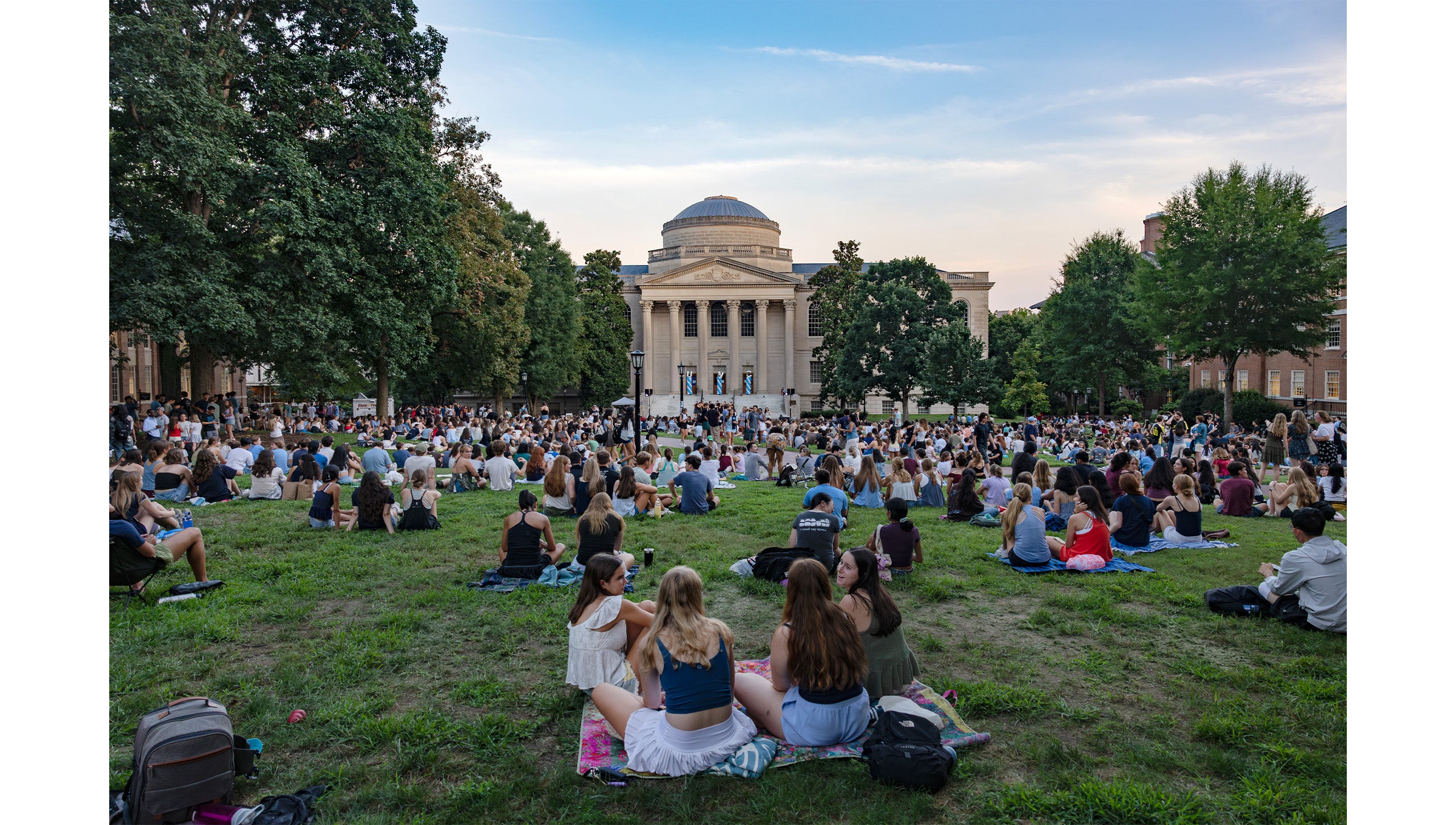 Wide-angle photo of students sitting on the lawn of Polk Place at the Sunset Serenade performance with Wilson Library seen in the background.