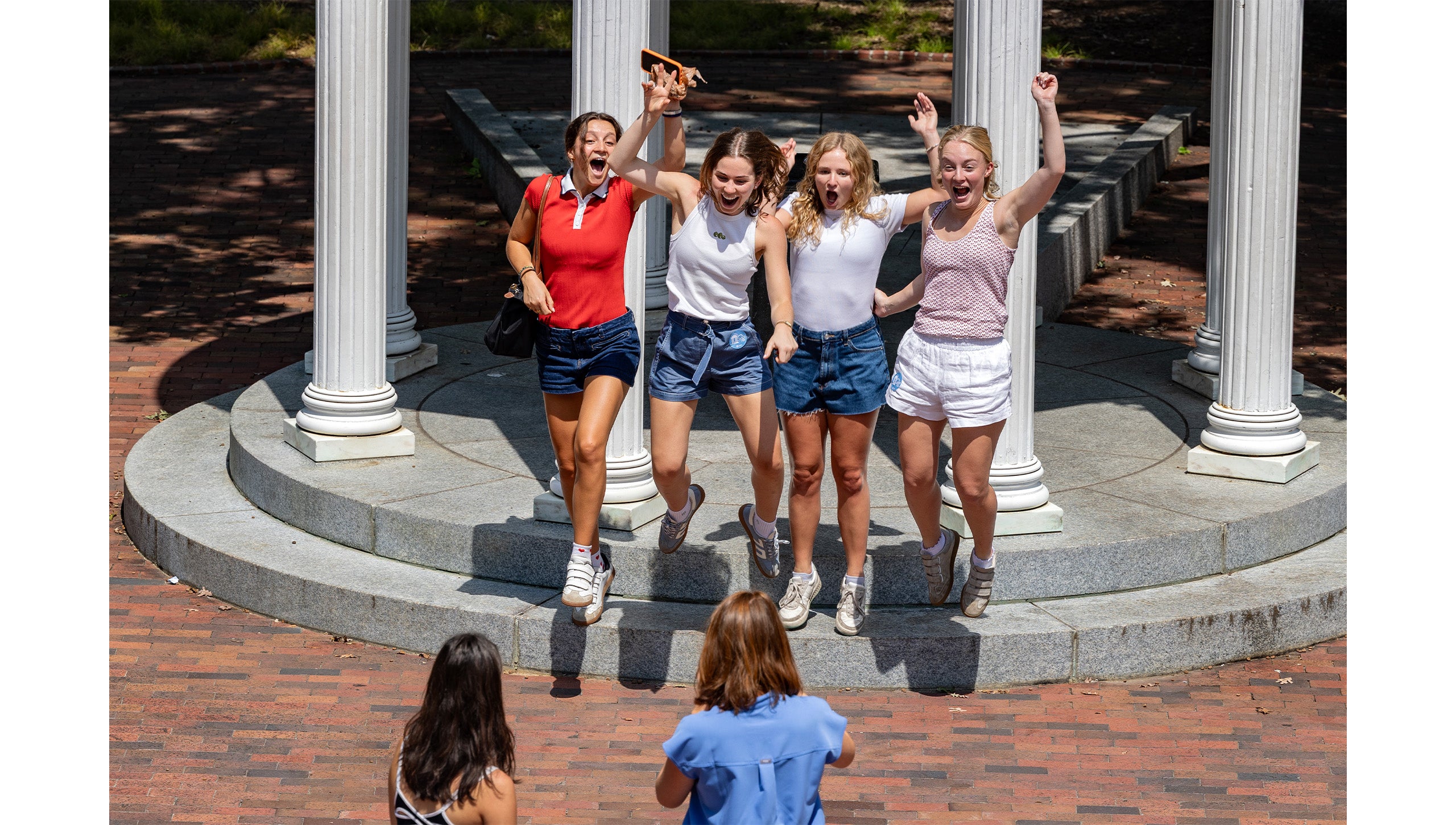 Group of four UNC-Chapel Hill students jumping together for a photo in front of the Old Well.