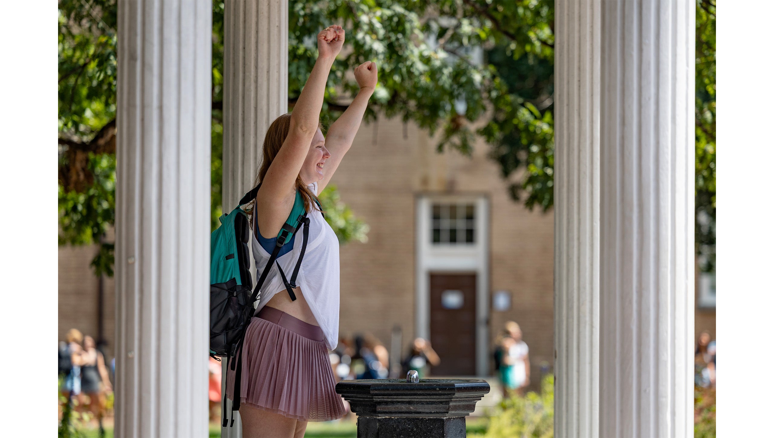Side angle of a UNC-Chapel Hill student putting her arms up in the air in celebration while standing behind the fountain of the Old Well and posing for a picture.