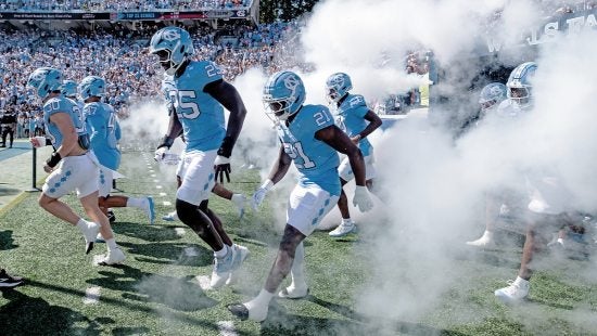 UNC football players running out the tunnel at Kenan Stadium onto the field through white smoke.
