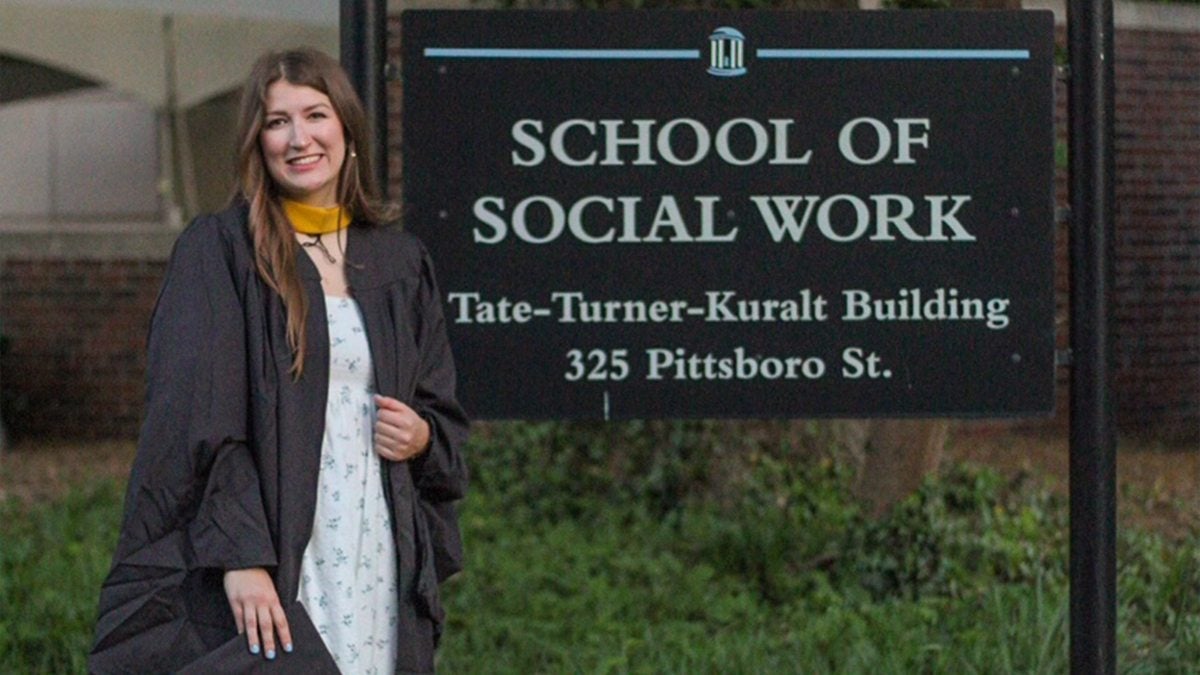 Hannah Collins in Black graduation regalia smiling and posing for a photo by the UNC-Chapel Hill branded building sign reading, 