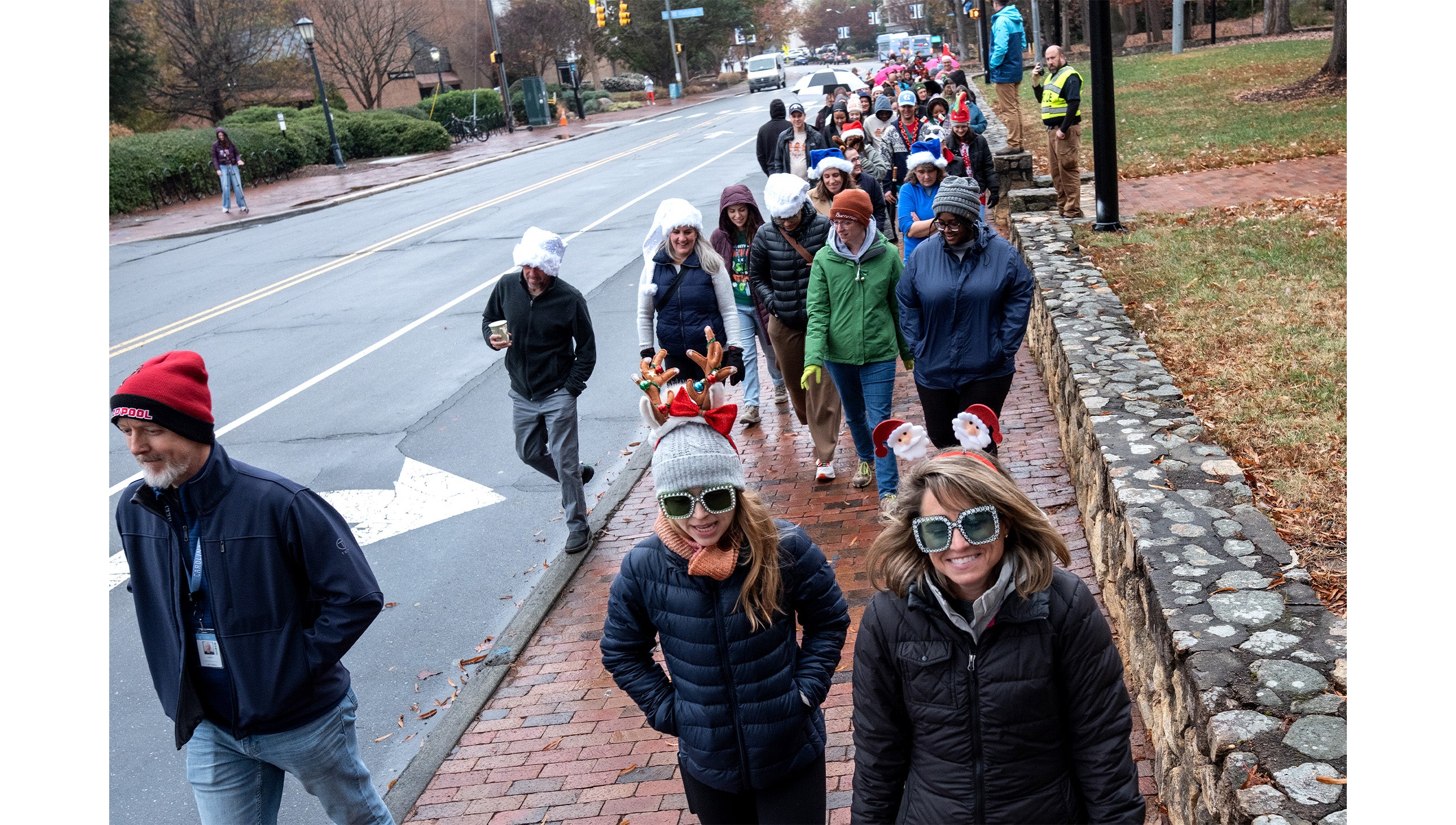 Long line of UNC-Chapel Hill employees in festive holiday costumes walking down a brick sidewalk aside South Road during the Jingle Bell Jog.