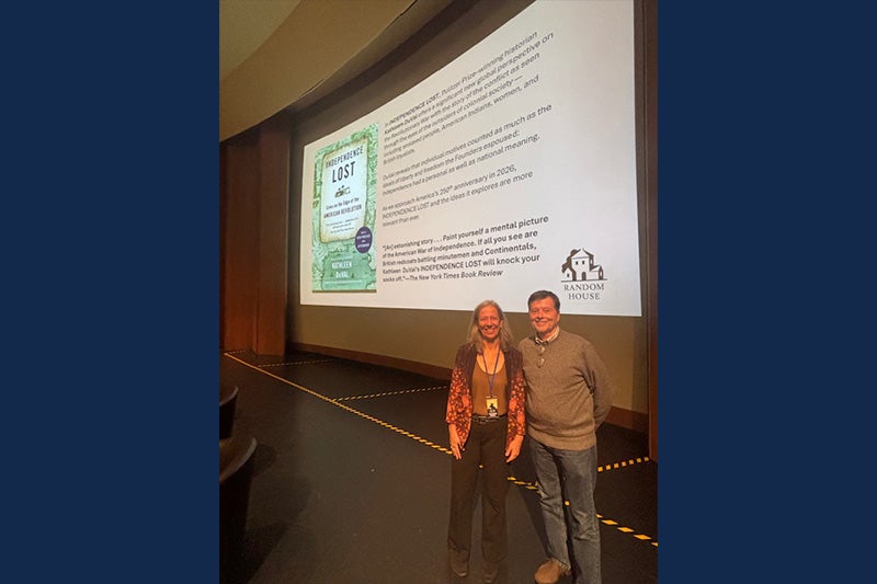 Kathleen DuVal standing next to Ken Burns on a stage with a projector displaying the cover of one of DuVal's books, "Independence Lost."