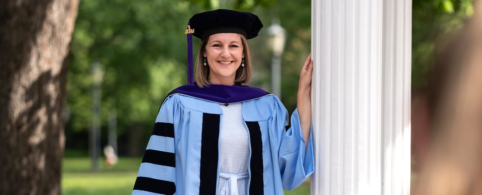 Brittany Akers posing for a graduation photo in Carolina Blue and black regalia while standing against one of the pillars of the Old Well on the campus of UNC-Chapel Hill.