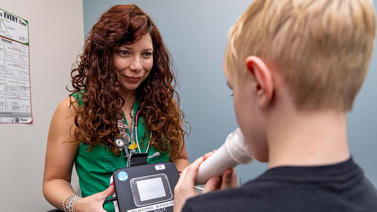 Michelle Hernandez watches as a young boy blows into a medical device during a breathing test in a clinic setting.