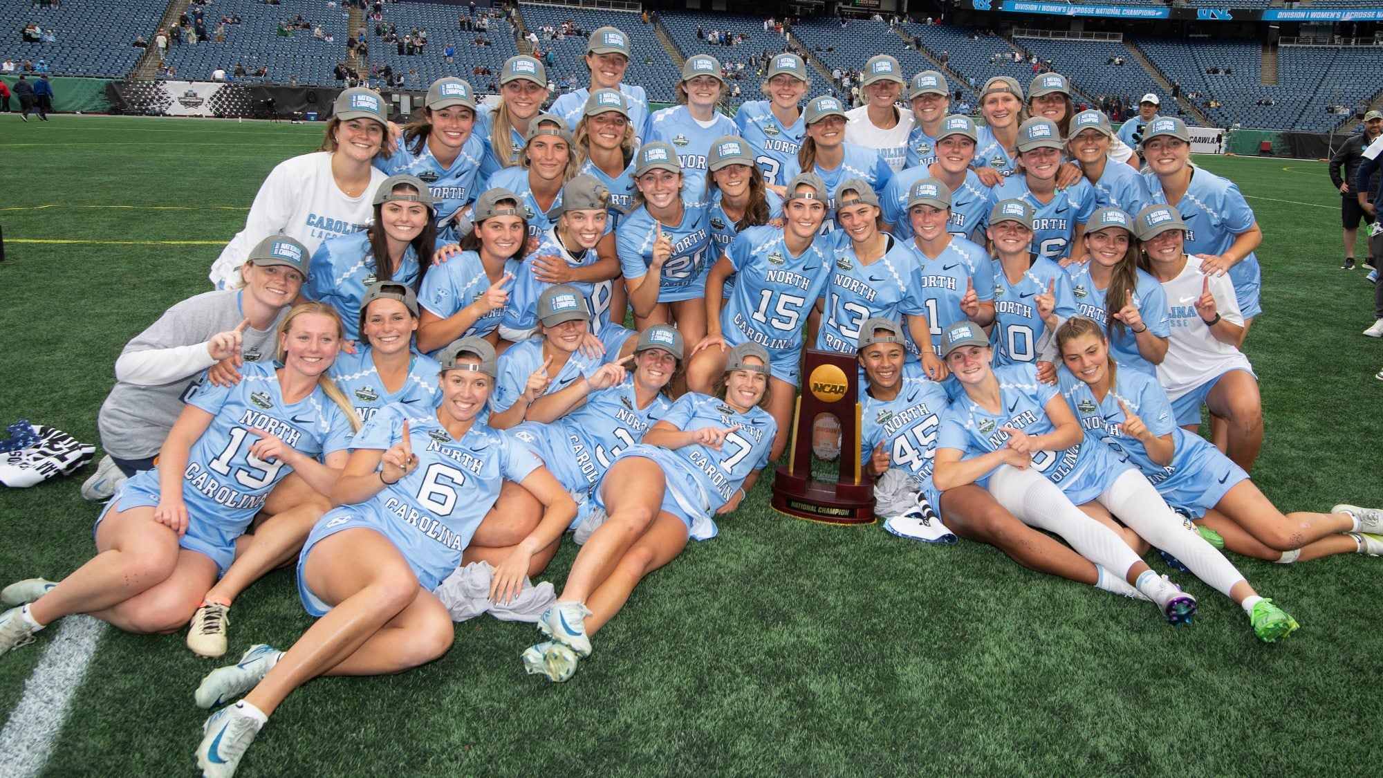 UNC women's lacrosse team celebrating and taking team photo on field after game with national title trophy.