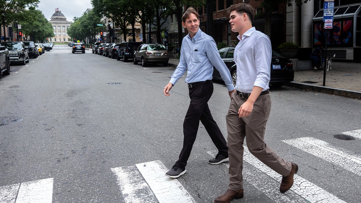Reece Hawk and Andrew Stewart talking as they cross Fayetteville Street at a crosswalk in downtown Raleigh. The North Carolina State Capitol is seen in the distance.