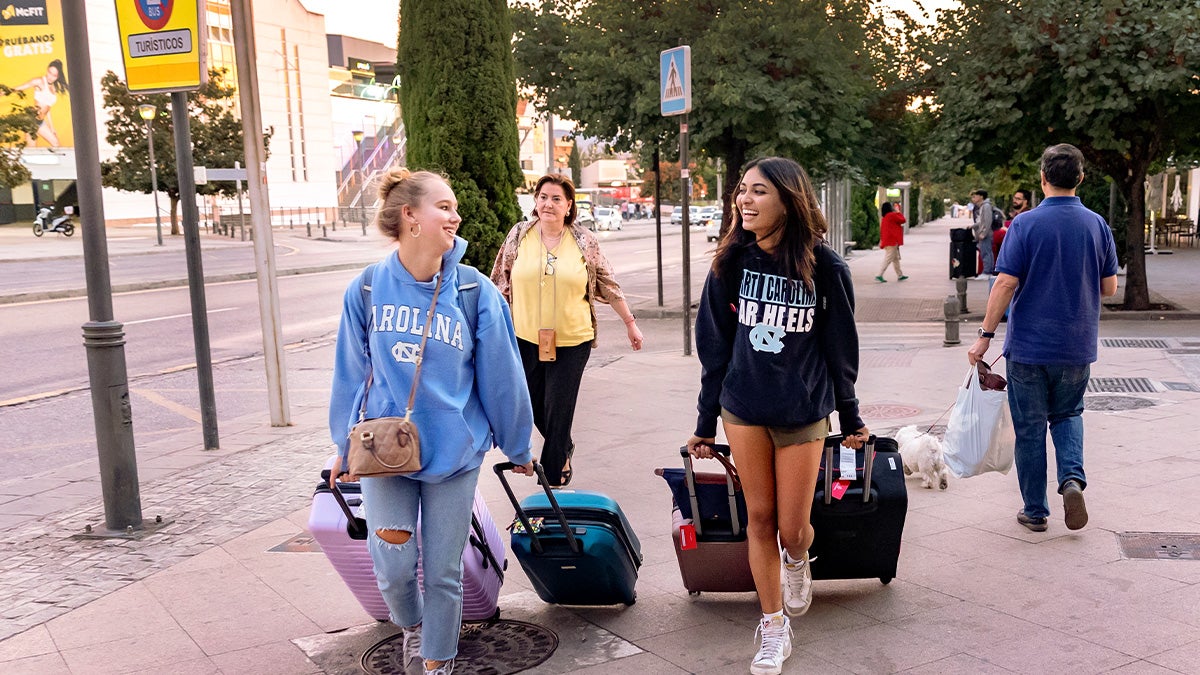 Two students wearing UNC-Chapel Hill hoodies carrying suitcases down a sidewalk in Granada, Spain.