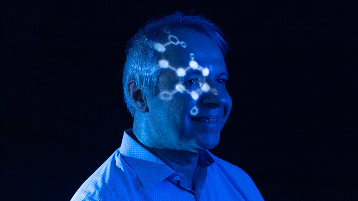 Alex Tropsha poses for a photo, his face illuminated by projected atoms and bathed in a cool blue hue.