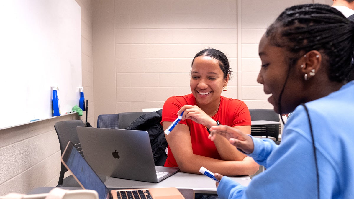 A student tutor sitting a desk with a laptop tutoring a student who also has her laptop out.