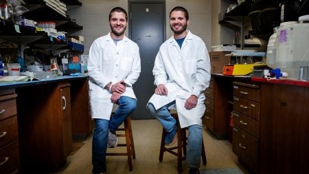Two twins, Ryan and Brandon Mouery, wearing lab coats and posing for a portrait while sitting on stools in a lab.