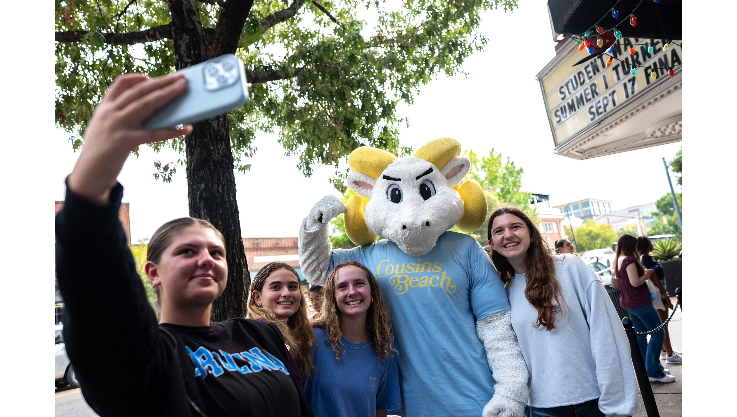 Four UNC-Chapel Hill students taking a selfie with Rameses, UNC-Chapel Hill's ram mascot, outside the Varsity Theatre on the sidewalk of Franklin Street.