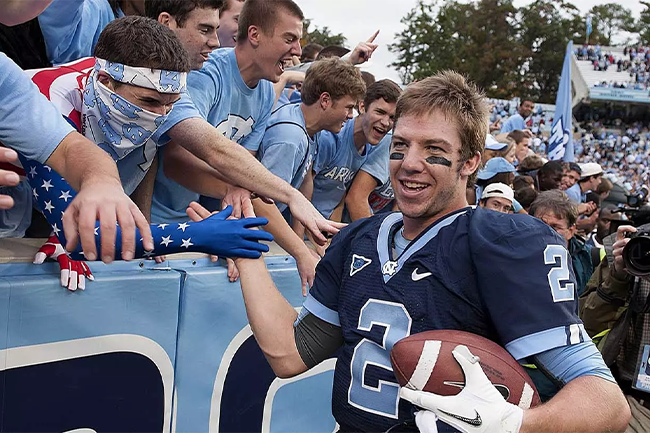 Bryn Renner as a Carolina student wearing his football uniform, high-fives students as they cheer him on during a game.