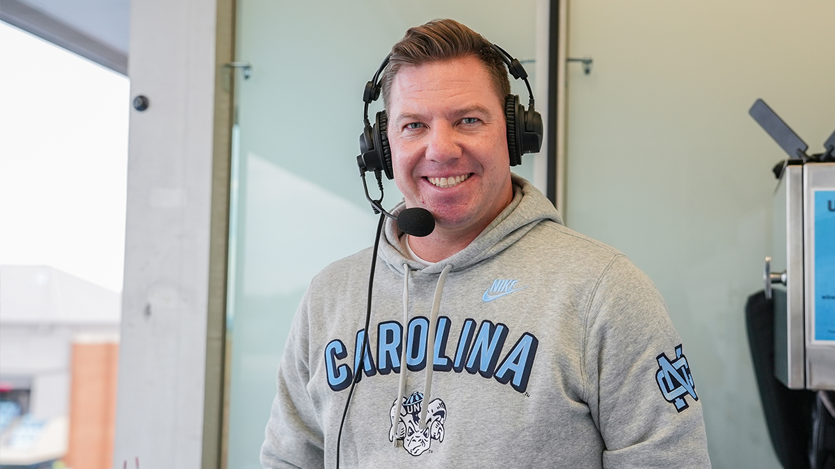 A portrait of Bryan Renner smiling at the camera wearing a Carolina hoodie and a pair of over the ear headphones with a mic attachment.