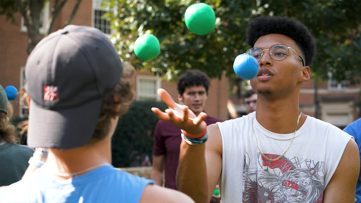 One student juggling a three colorful balls in front of another student - the ball float in the air in this photograph.