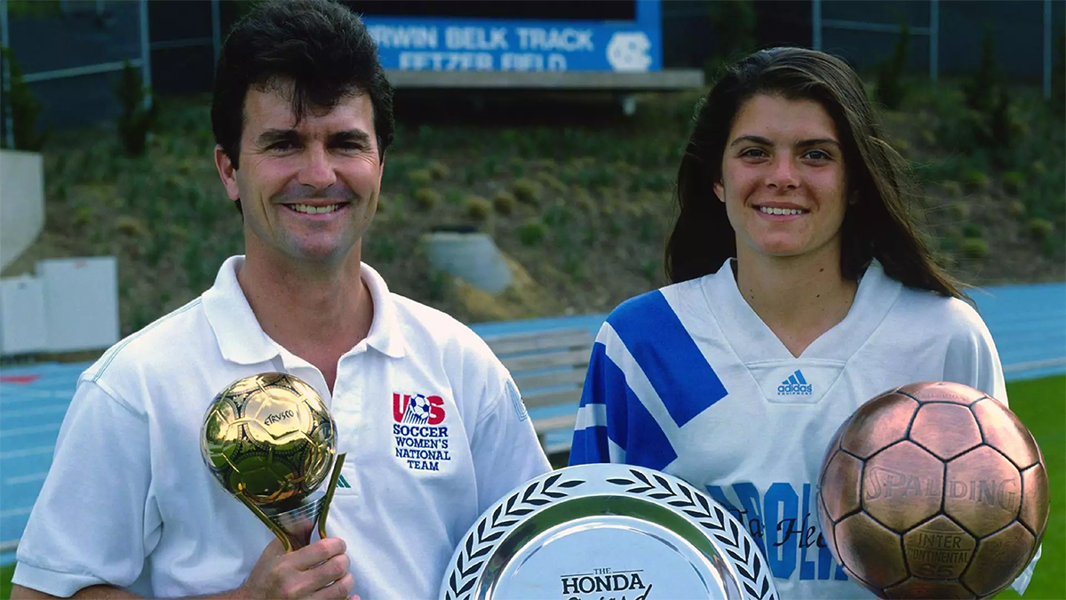 Anson Dorrance in a white U.S. Soccer collared shirt holding a gold World Cup trophy and Mia Hamm in her Carolina soccer uniform holding two trophies for soccer awards she won.