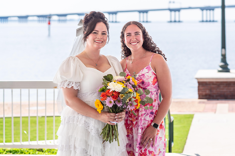 Lindsey Meier (Left) bride in a white dress holds a colorful bouquet and stands beside Maddie Hoffman (Right), who is wearing a pink floral dress. They smile together outdoors with a waterfront and bridge in the background.