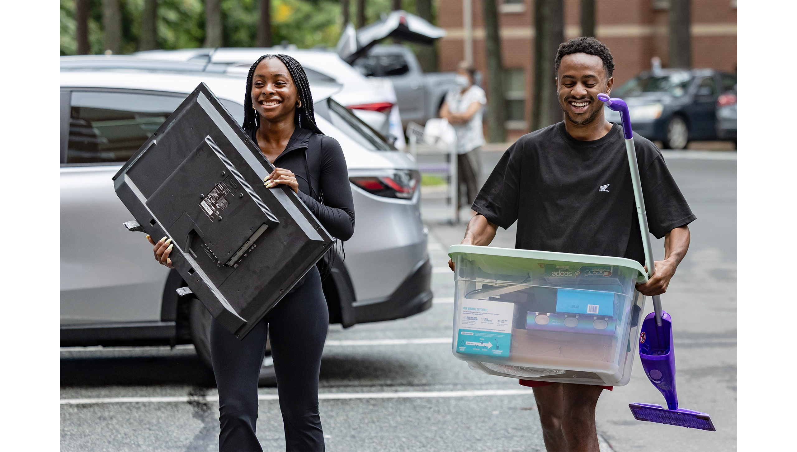 Two people holding a TV and a plastic crate in the parking lot.
