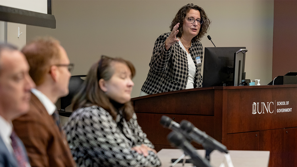 A woman at a podium, speaking into a mic, wit attendees of event in foreground.