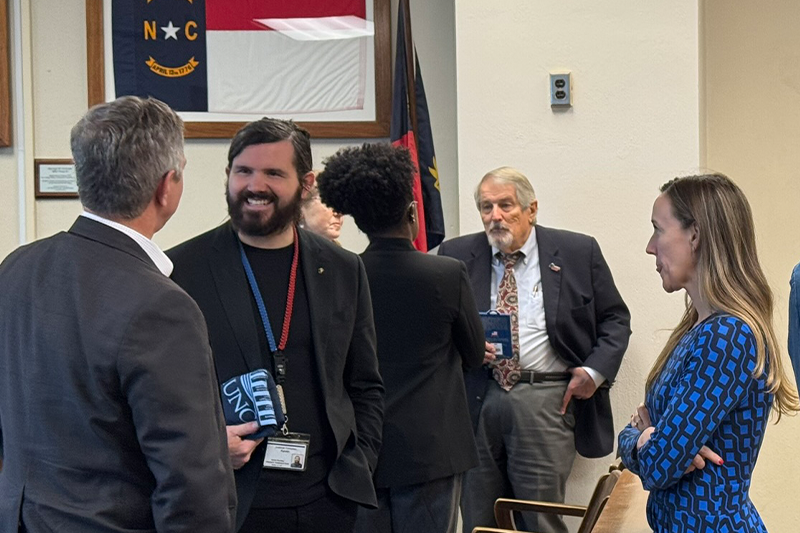 A group of six people smiling and talking in a room with a North Carolina flag and state seal displayed on the wall. Four of the individuals are standing, while one is seated, all engaged in conversation.