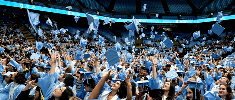 Graduates in Carolina blue gowns celebrating at winter commencement, tossing their caps into the air inside a crowded arena.